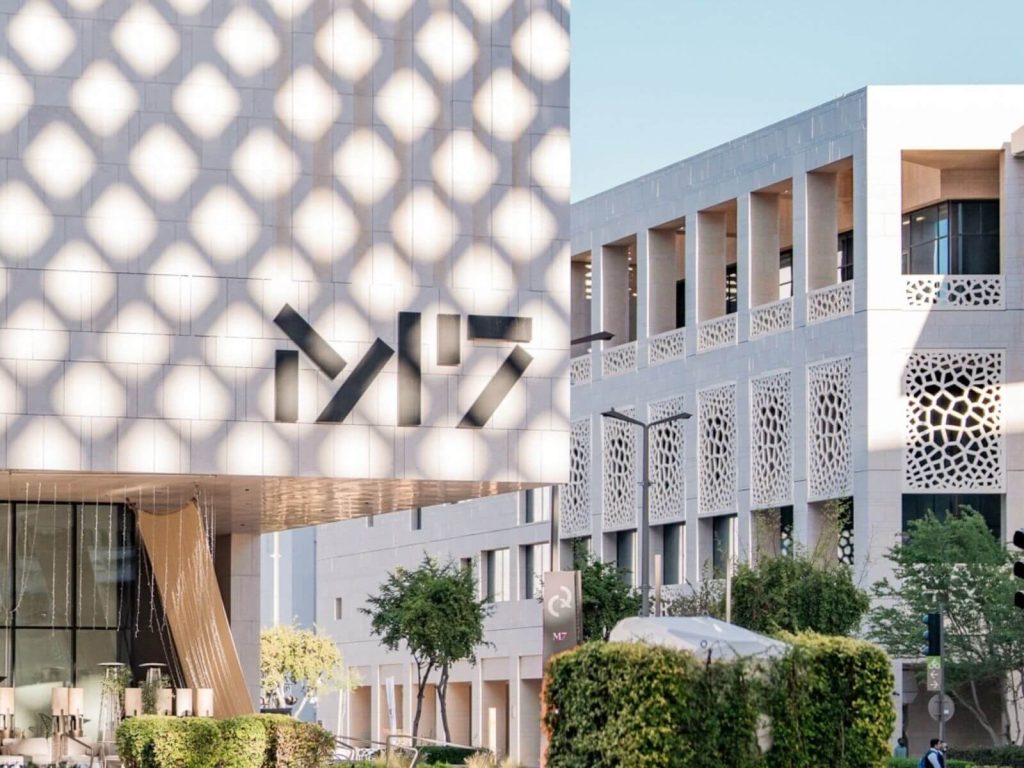 Exterior architectural shot of the M7 creative hub in Doha, Qatar, showing modern white buildings with traditional geometric shadow patterns and greenery.