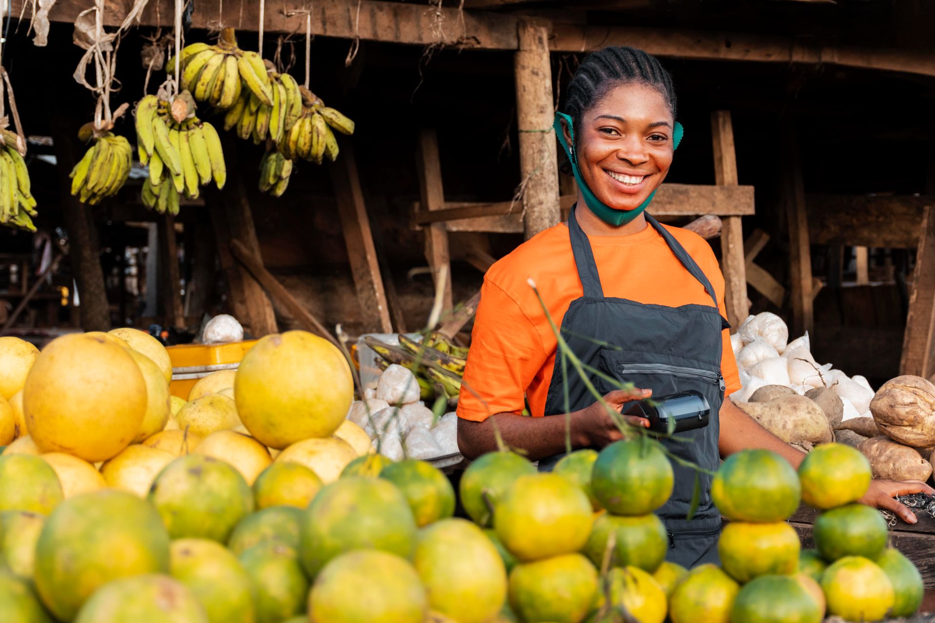 A smiling young African female agricultural entrepreneur holding a digital POS machine at a vibrant fresh produce market, representing modern agri-tech and food security in Africa.