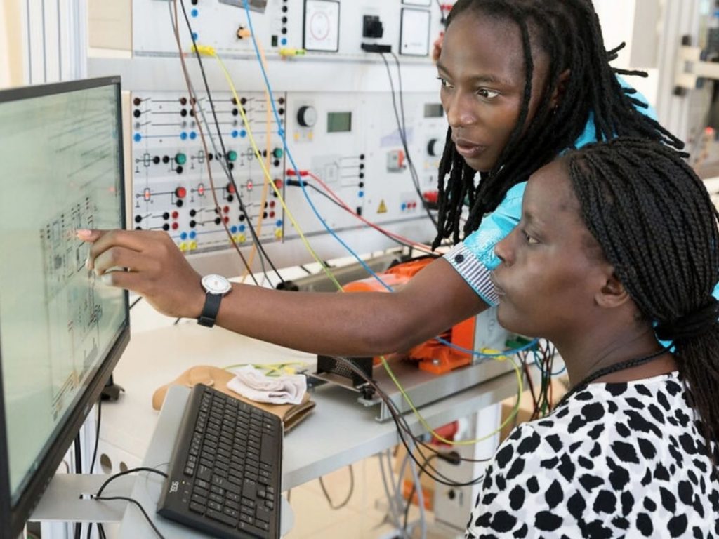Two young African female tech professionals analyzing complex systems data on a computer monitor in a hardware engineering lab, representing the development of predictive infrastructure for food security.
