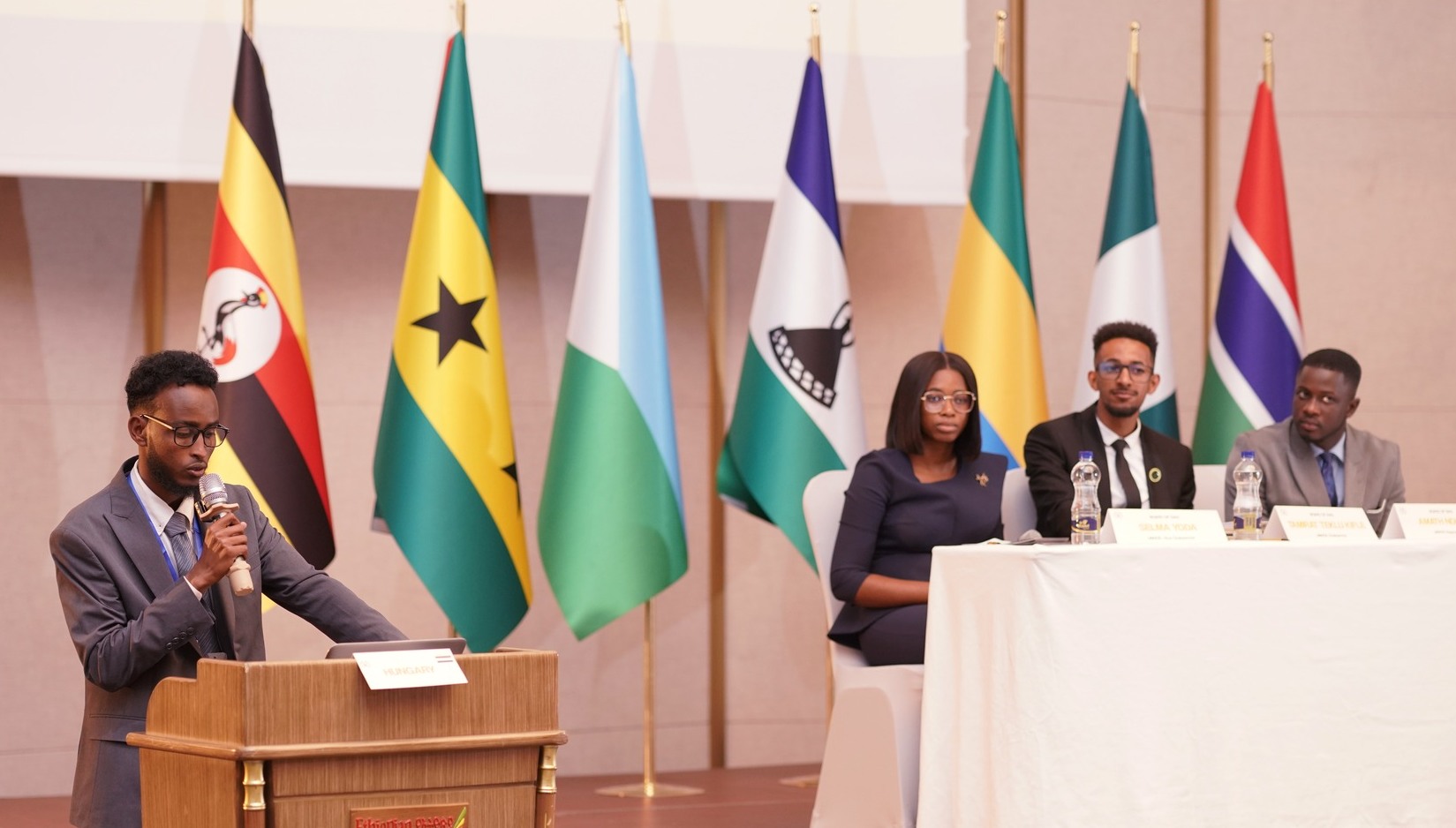 Young African diplomat speaking at a podium with a panel of youth leaders and African national flags in the background.