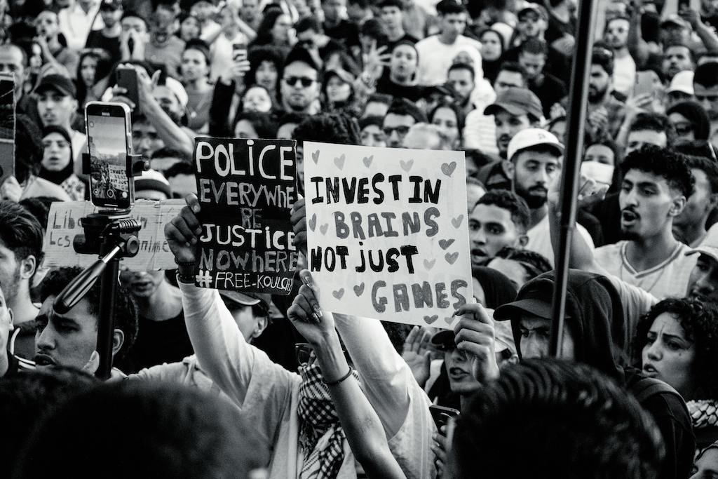 A dense crowd of protestors in a city, with two prominent hand-written signs reading 'Police Everywhere Justice Nowhere' and 'Invest in Brains Not Just Games,' symbolizing the demand for merit-based governance and social justice