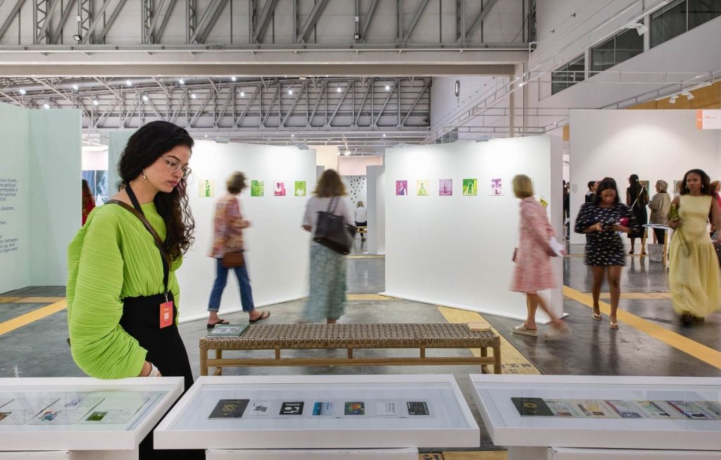 A wide-angle view of a bustling modern African art fair with visitors viewing an exhibition about identity and passports, highlighting the scale of Africa's creative industry.