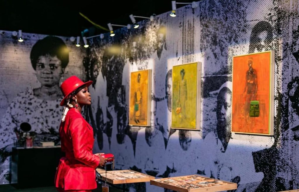 A stylish young African woman in a vibrant red suit viewing contemporary paintings at a modern art exhibition, representing the growth of Africa's creative economy.