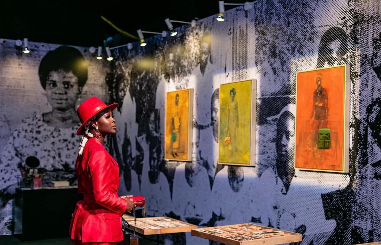 A stylish young African woman in a vibrant red suit viewing contemporary paintings at a modern art exhibition, representing the growth of Africa's creative economy.