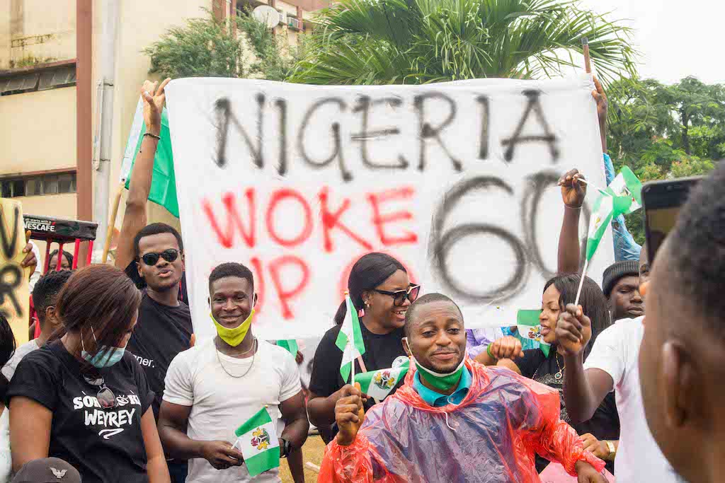 A vibrant group of young Nigerian protestors holding a large white banner that reads 'NIGERIA WOKE UP @ 60' while waving national flags, representing the grassroots movement for social justice and institutional awareness in Africa