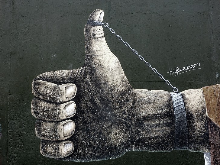 A street art mural of a large hand giving a thumbs-up gesture while the thumb is shackled to the wrist by a metal chain, symbolizing the structural restraints on social progress and the weaponization of 'woke' rhetoric