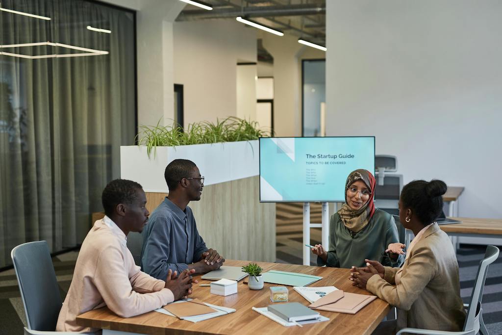 A diverse group of African professionals, including a woman in a hijab, collaborating in a modern office with a 'Startup Guide' presentation, symbolizing the economic competitive advantage of diversity in the global marketplace