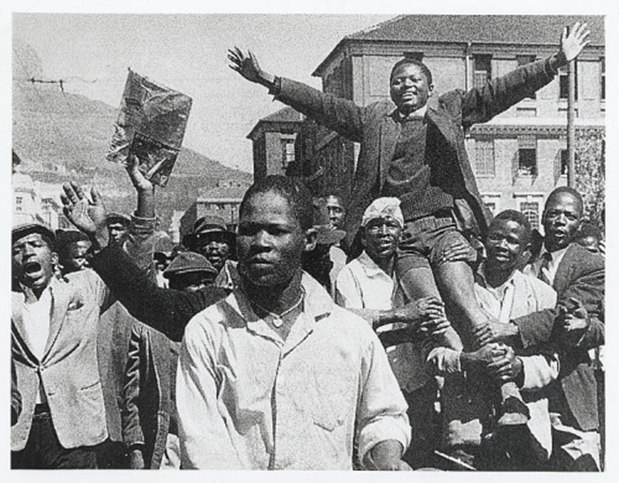 Black-and-white photo of the 1960 anti-Apartheid pass law protest in Cape Town. A crowd of Black South African men march in defiance against the laws that systematically excluded them from the economy.