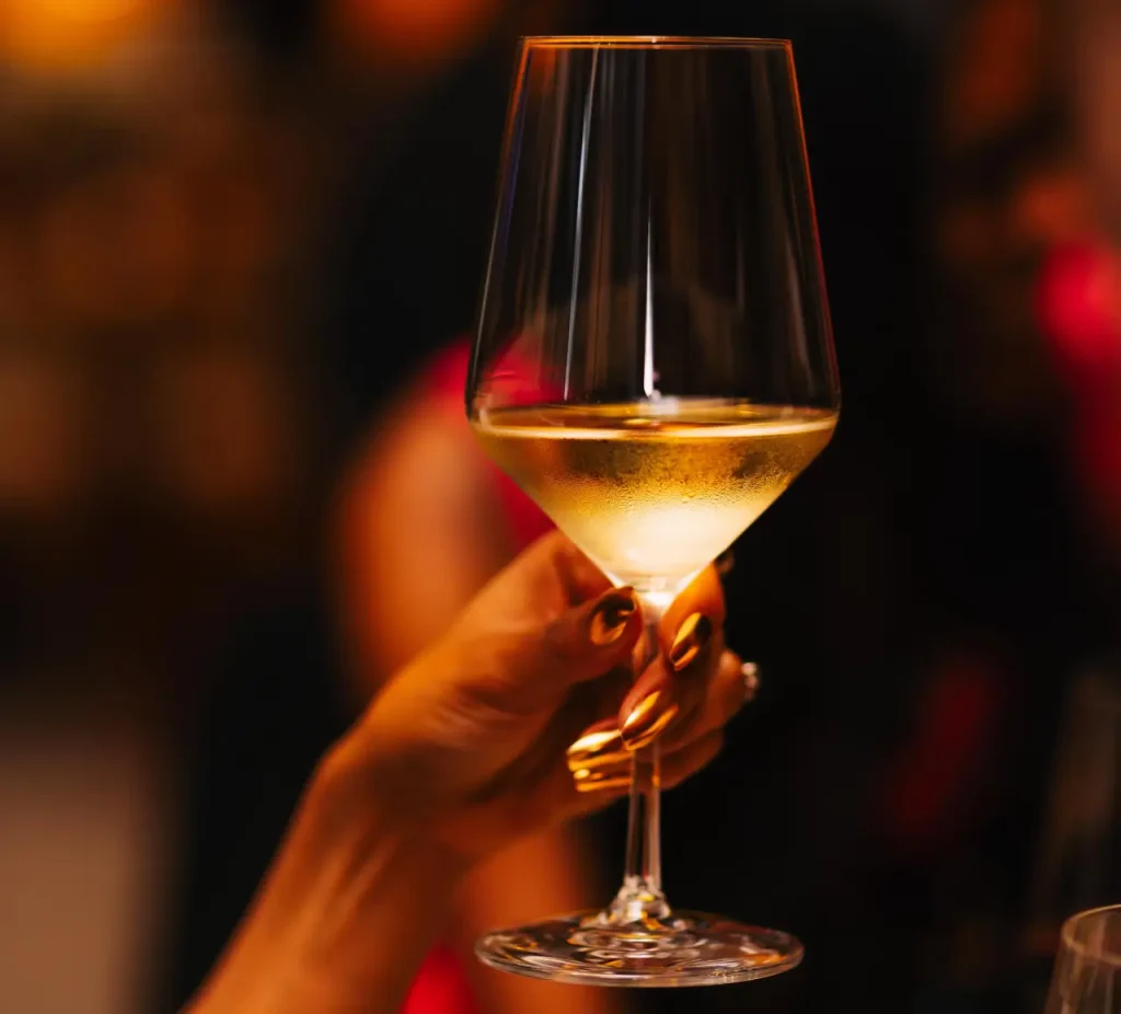 A hand holding a glass of white wine with a blurred, warm-toned background suggesting a sophisticated social event