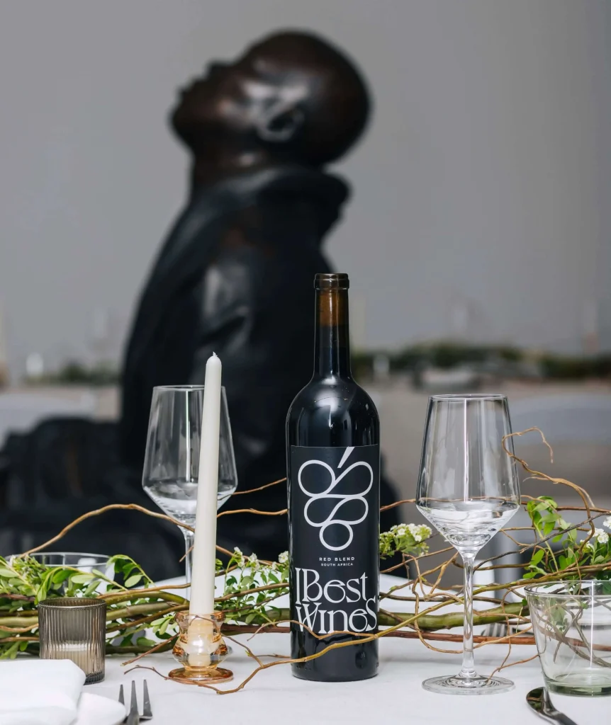 A bottle of IBest Wines Red Blend from South Africa on a white tablecloth, flanked by empty wine glasses, a candle, and green foliage, with an abstract dark sculpture in the blurred background.