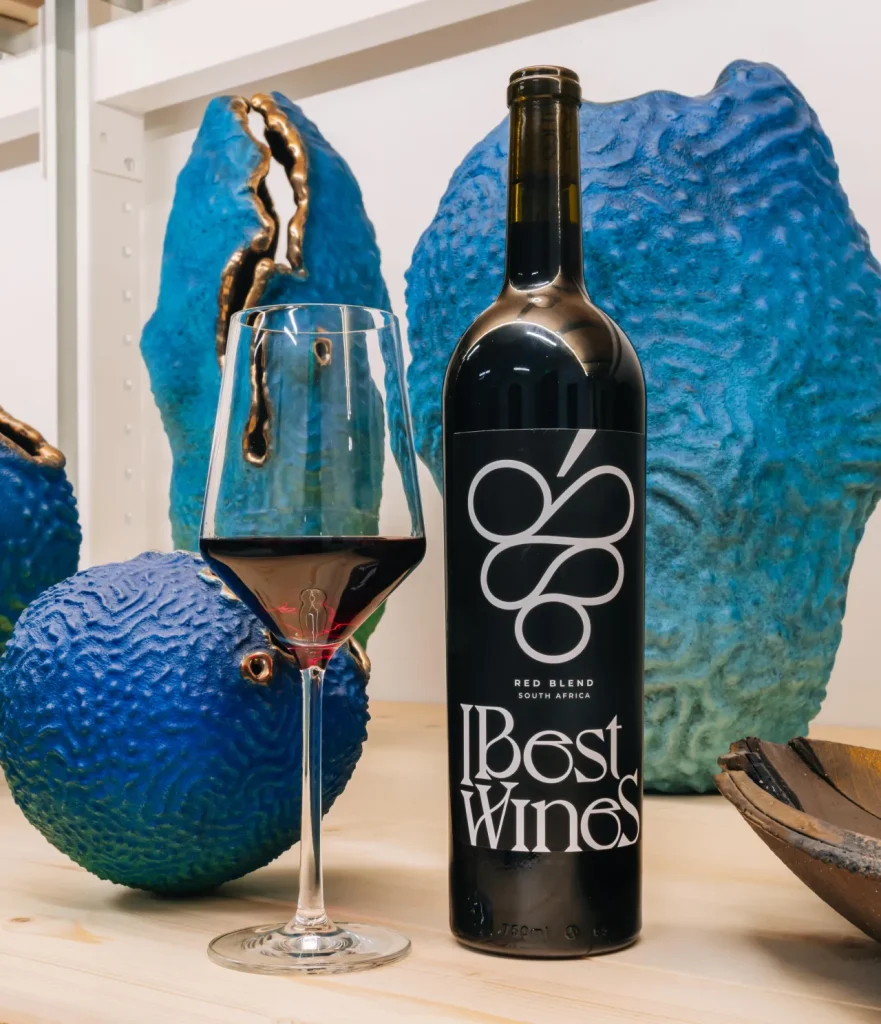 A bottle of IBest Wines Red Blend from South Africa and a glass of red wine, displayed on a wooden surface with vibrant blue contemporary African sculptures in the background