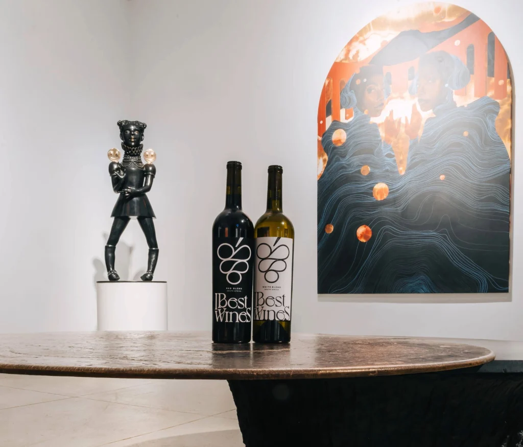 Two bottles of IBest Wines (Red Blend and White Blend) on a dark wooden table, with a contemporary dark sculpture on a pedestal and a vibrant arched wall painting in the background of an art gallery