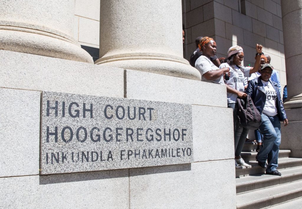 The High Court of South Africa's sign, with supporters on the steps behind it, symbolizing the public's scrutiny of the South African justice system in the Malema case.